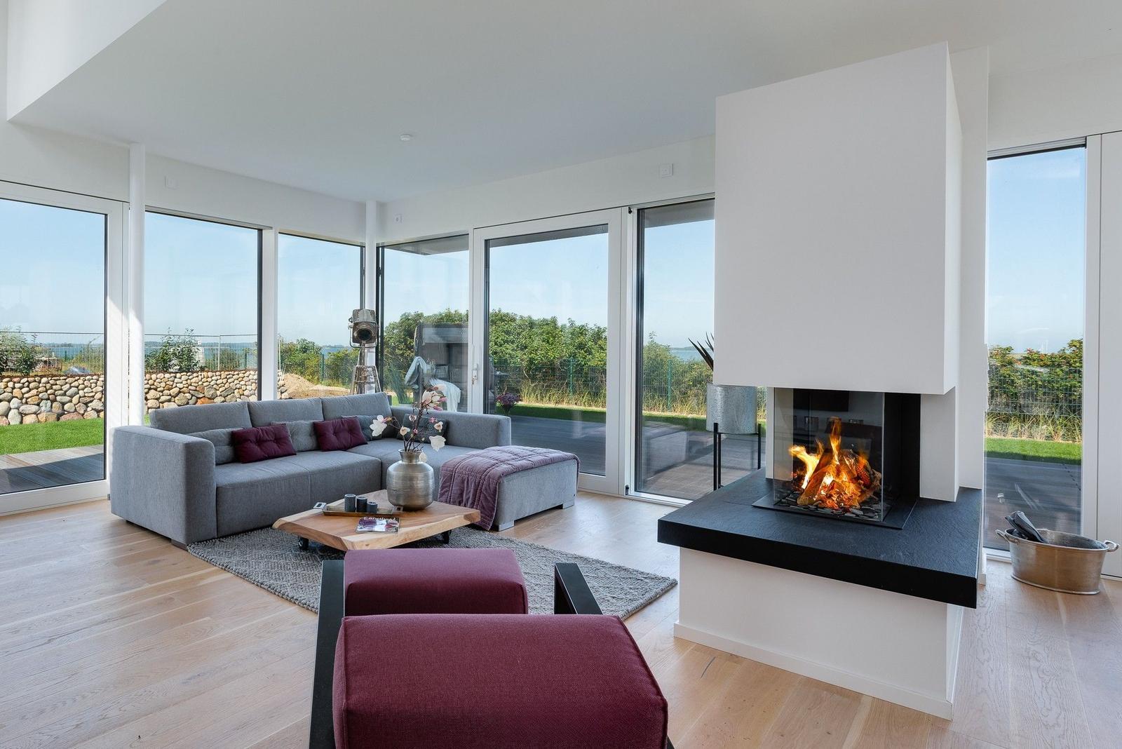 Ein Wohnzimmer mit Kamin, grauem Sofa und roten Sitzkissen. Große Fenster bieten Blick auf die Landschaft.