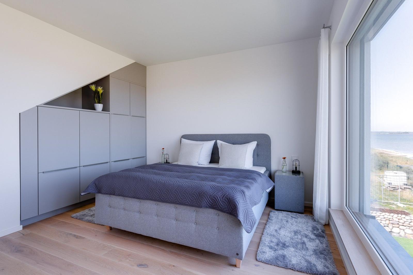 Schlafzimmer mit Bett, Kleiderschrank und großem Fenster mit Blick auf Wasser.