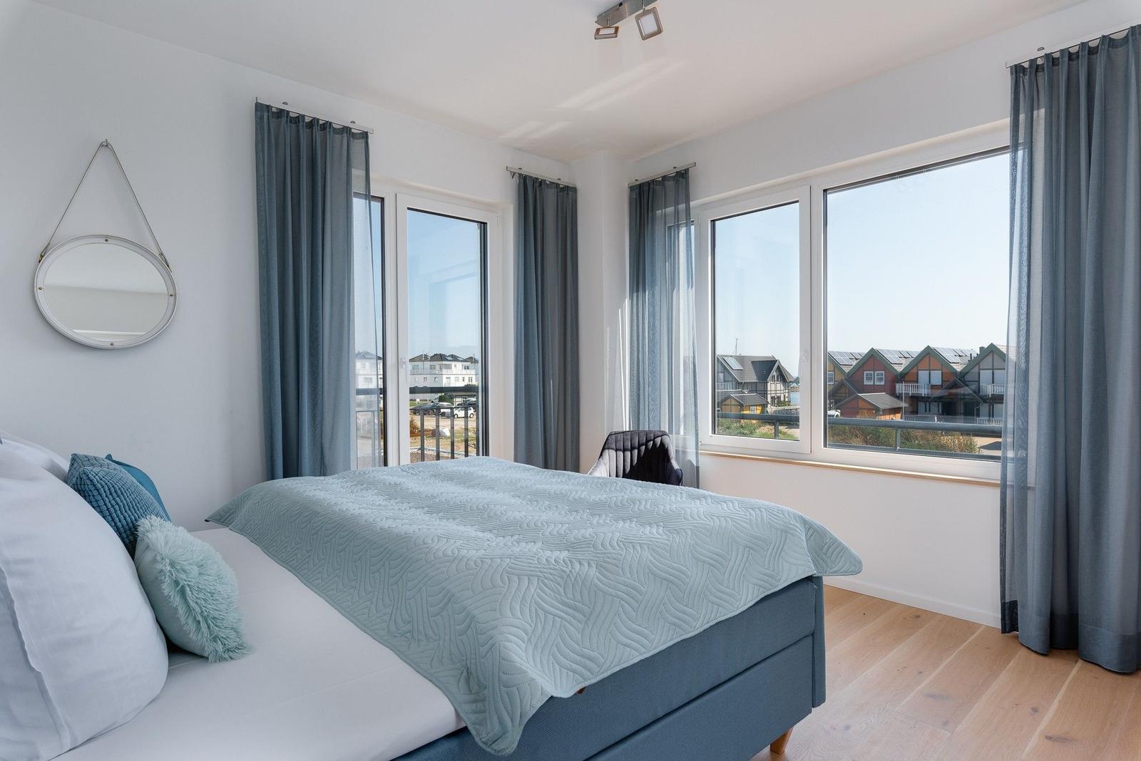 Schlafzimmer mit Bett, Fenstern und Blick auf Wohnhäuser.