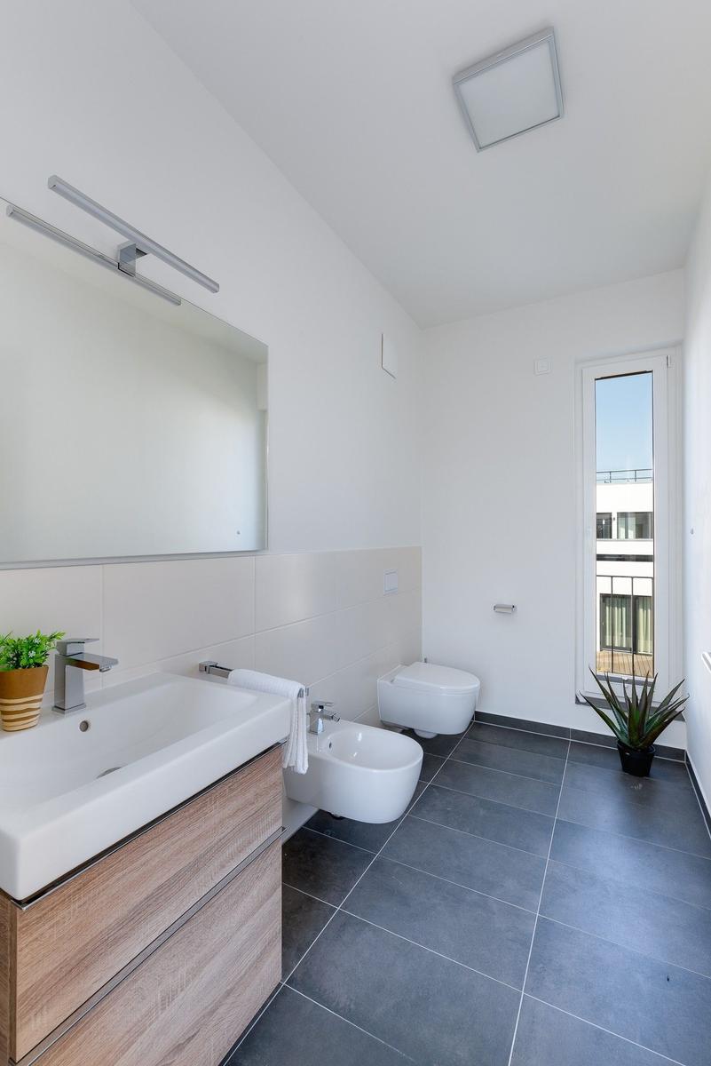 Badezimmer mit Waschbecken, Bidet, Toilette und Fenster. Holzunterschrank und Pflanzen.