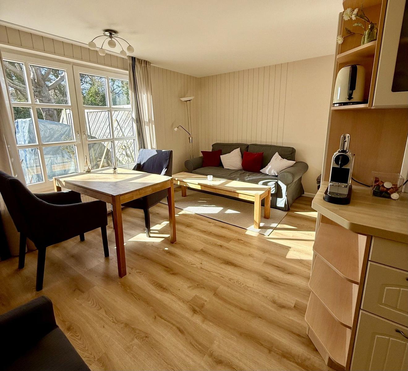 Wohn- und Essbereich mit Sofa, Tisch und Küchenzeile. Holzboden und große Fenster.