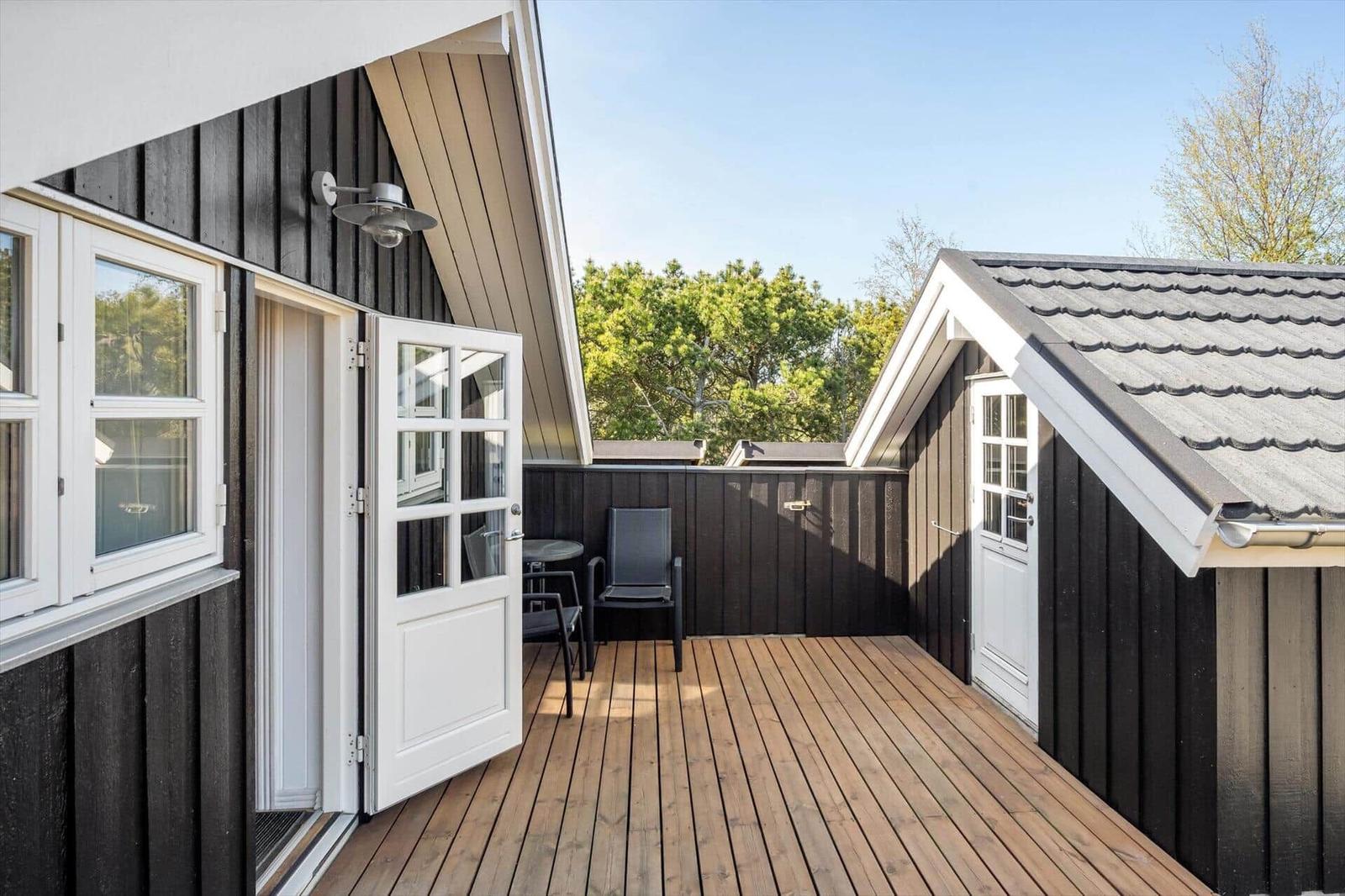 Deck with wooden floor, chair, and table. Black wood walls and white doors. View of trees.