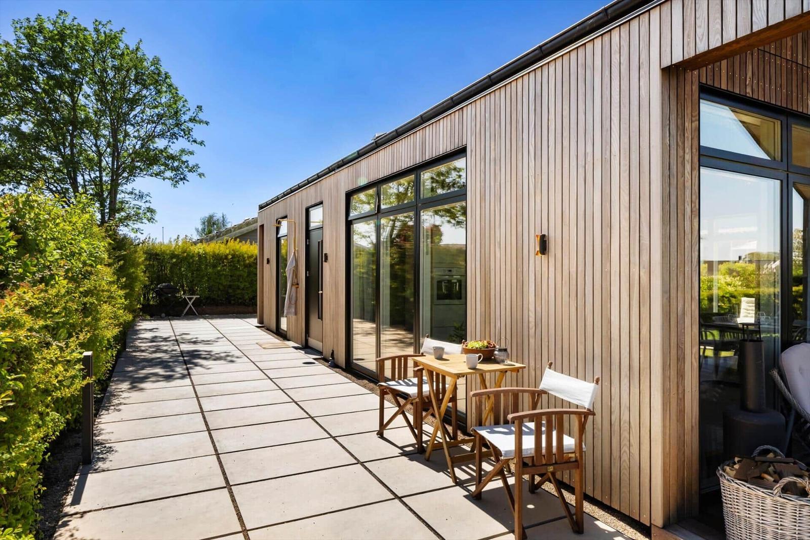 Terrace with wooden table and chairs beside modern wooden house with large windows.