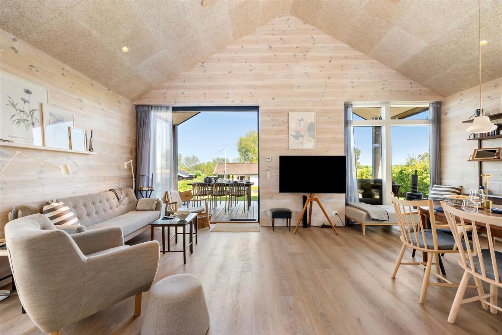 Living area with sofa, armchairs, TV, and dining area. Wooden walls and large windows.