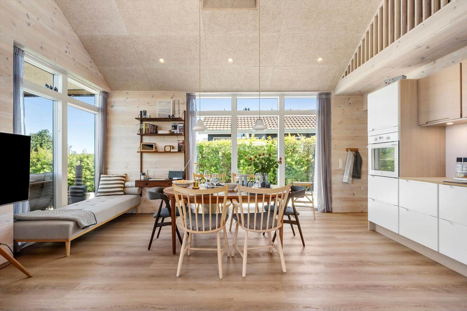Living kitchen with dining table, sofa, and large windows. Wooden floor and walls.