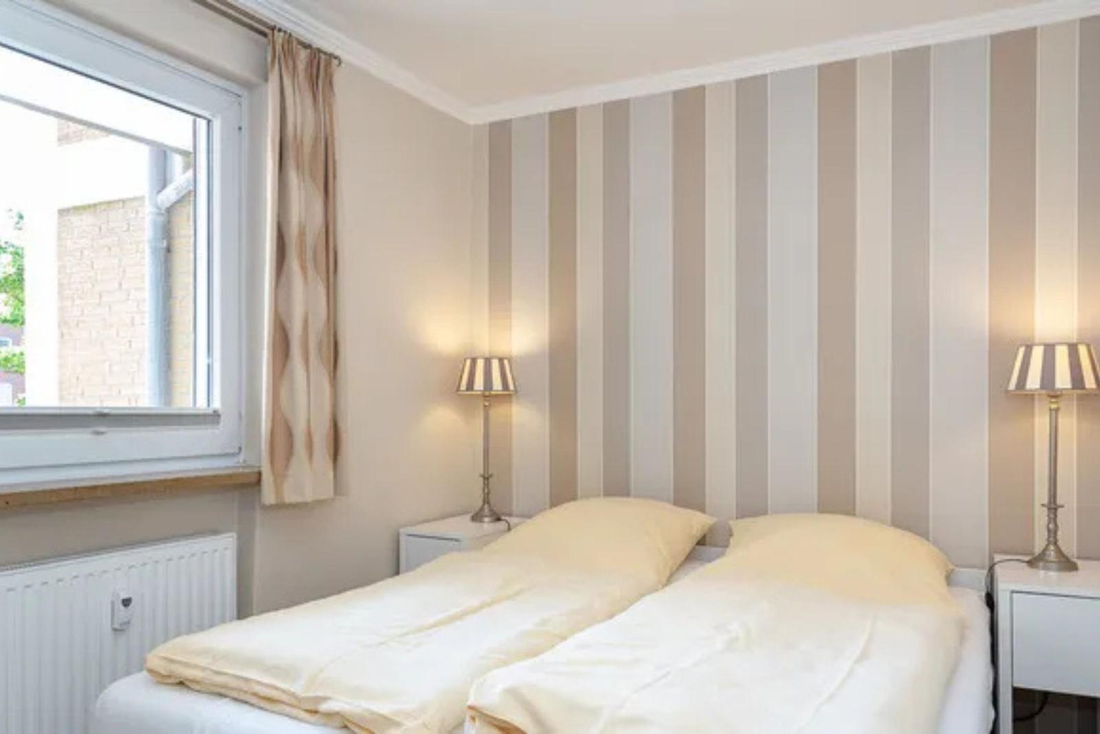 Schlafzimmer mit Doppelbett, Streifenwand, zwei Nachttischen und Fenster mit Vorhang.