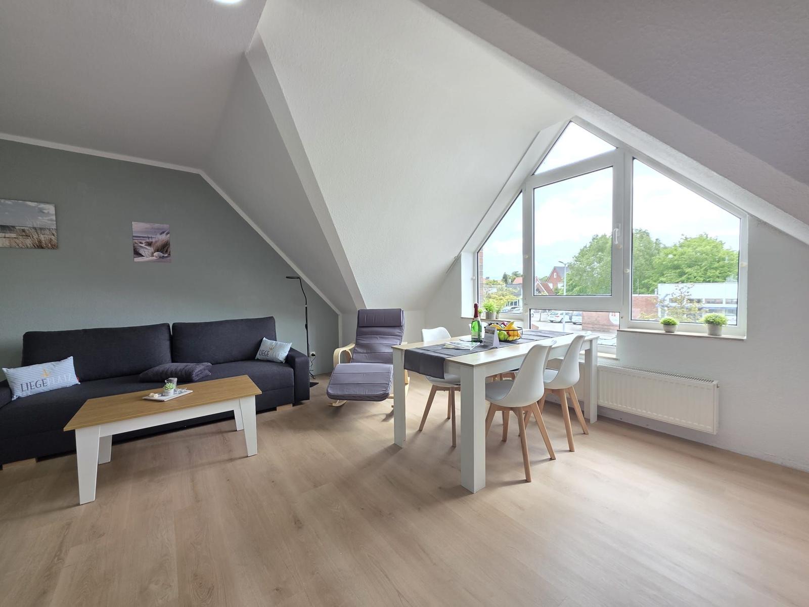 Living and dining area with sofa, table, and windows. Wooden floor and sloped ceiling.