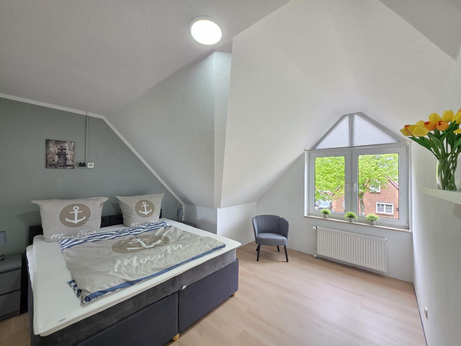 Bedroom with sloped ceiling, anchor-themed bed, armchair, and window view.