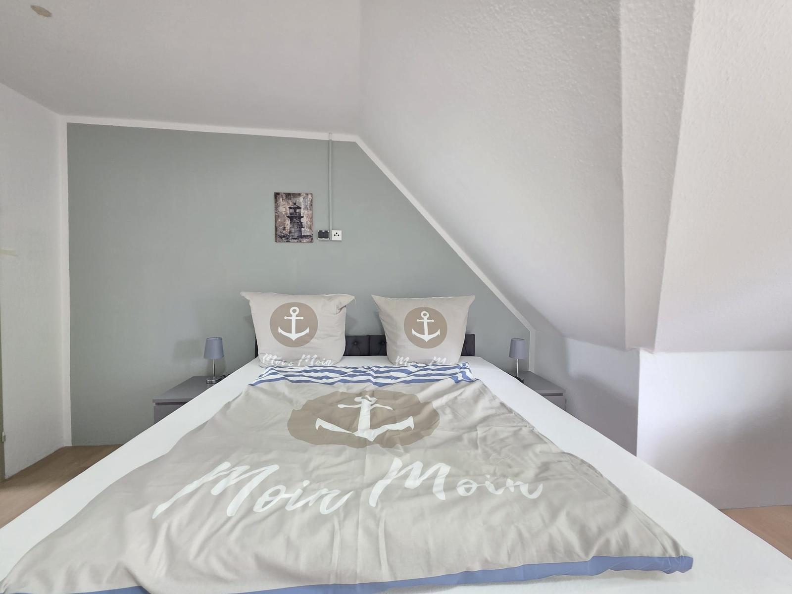 Bedroom with bed, pillows, and blanket featuring anchor motif. A lighthouse picture hangs on the wall.