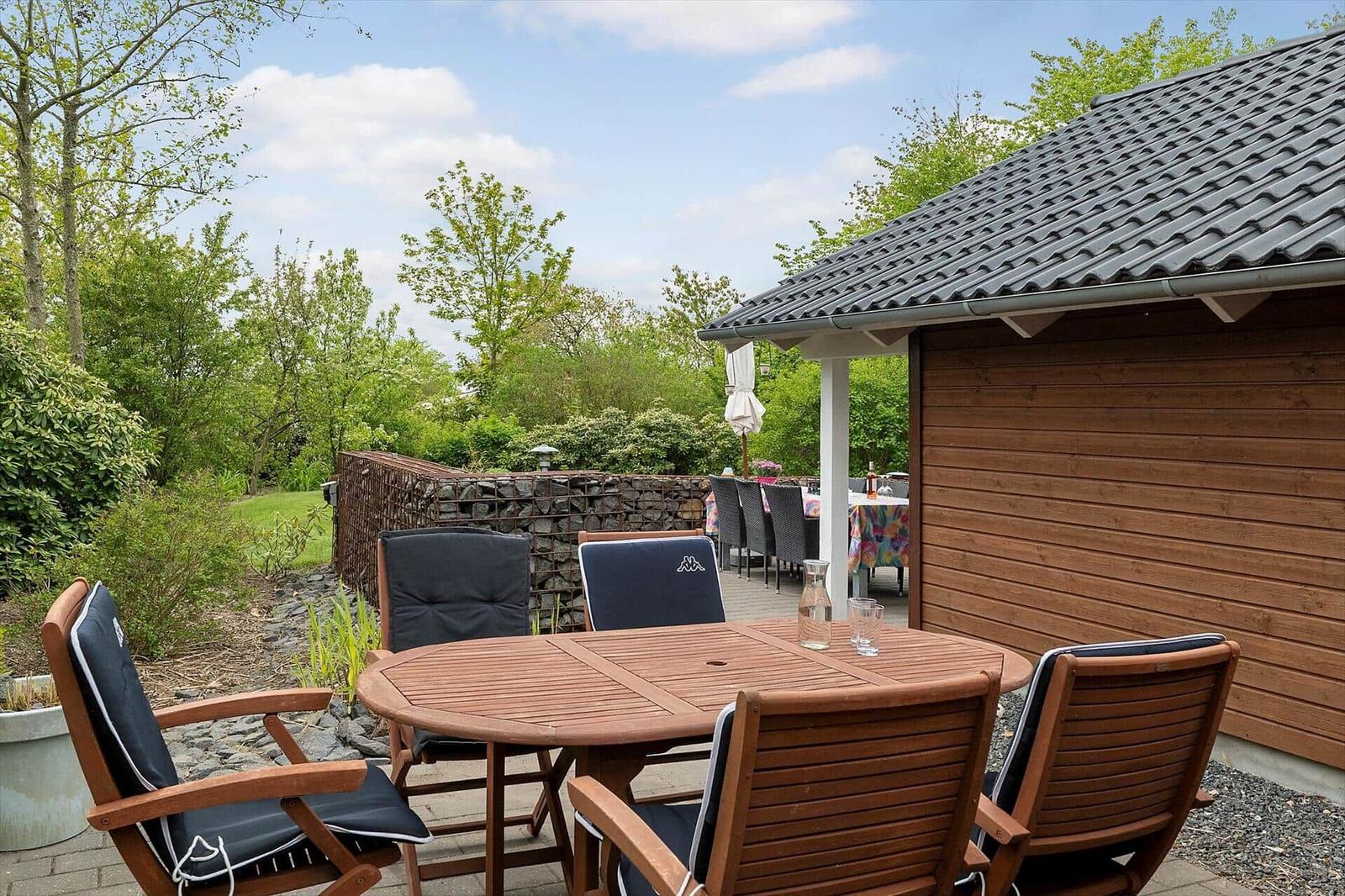 Terrace with wooden table and chairs beside a wooden house. Background: green areas and stone wall.