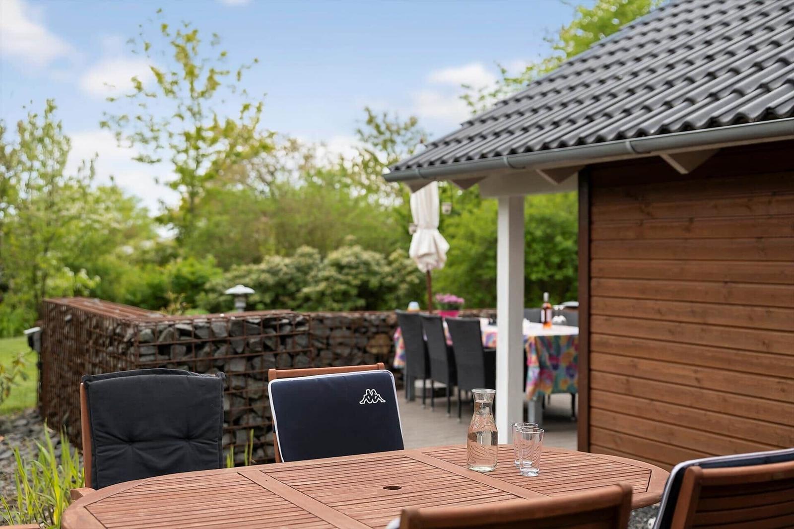 Wooden table with chairs in garden next to wooden house and stone wall.