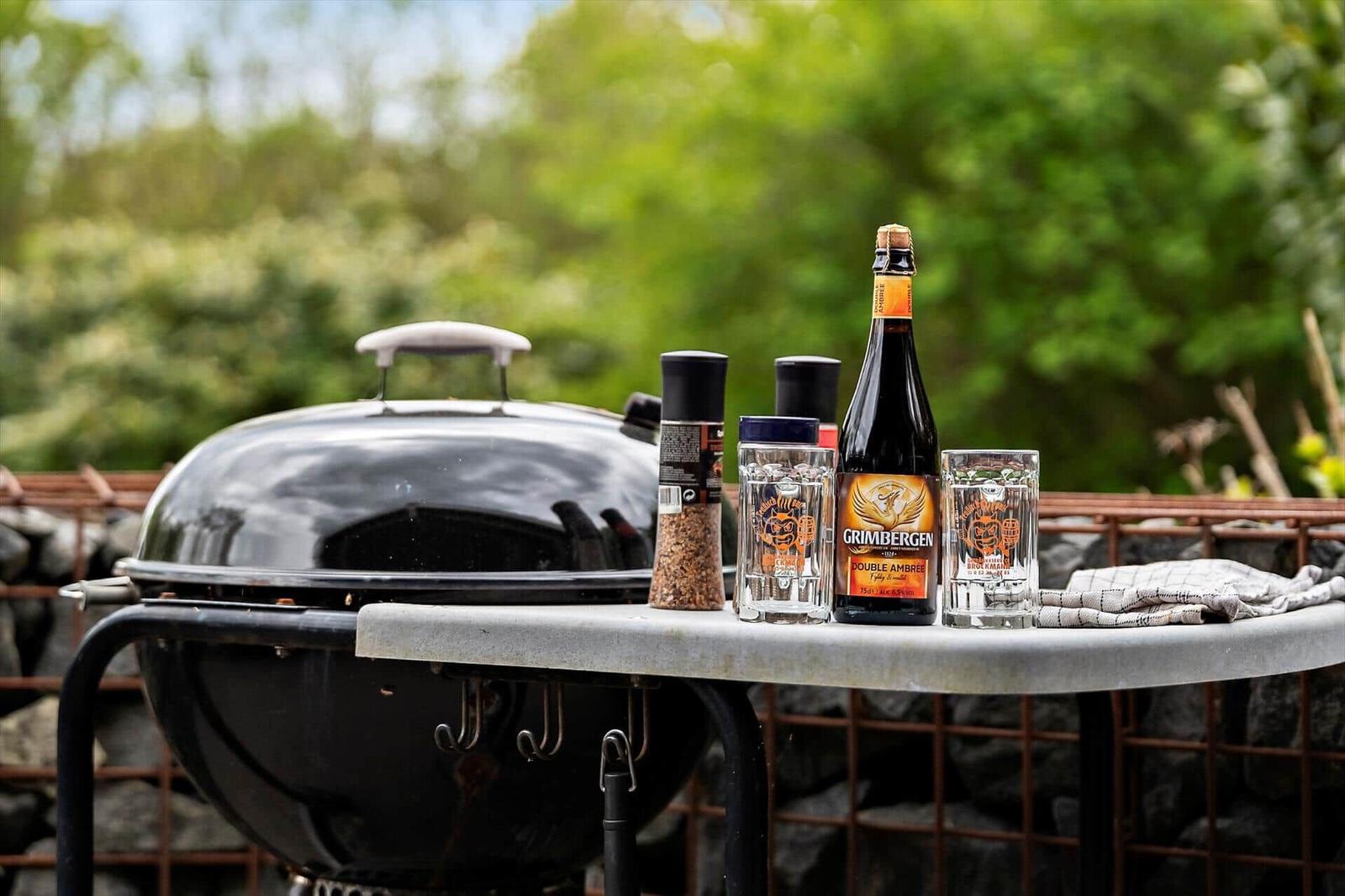 Closed grill with beer, glasses, and spice on table outdoors.