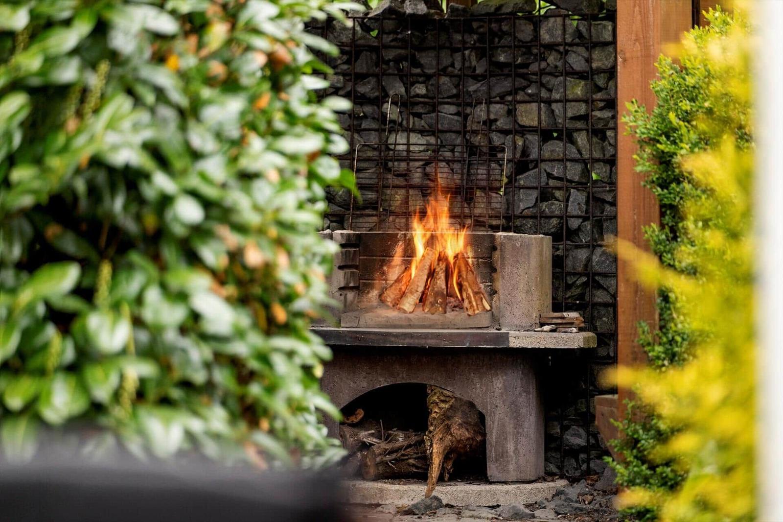 Outdoor area with burning wood stove, surrounded by plants and stone wall.