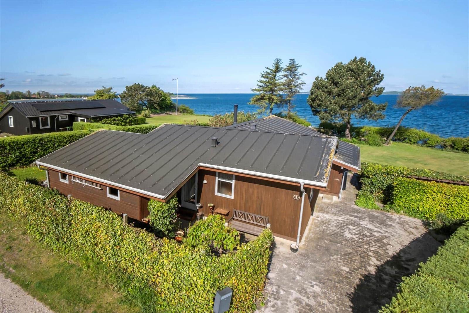 A house with wooden facade and gray roof, surrounded by greenery and overlooking the sea.