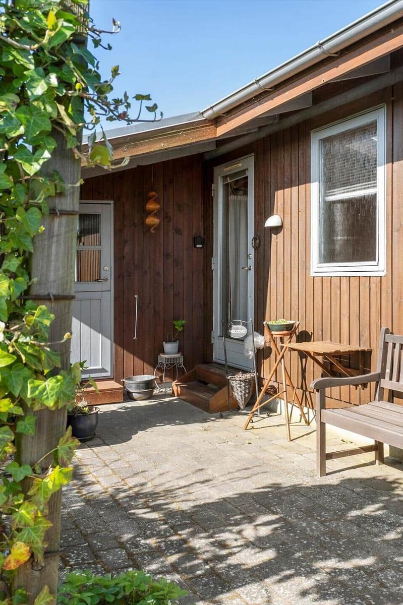 House with wooden siding, patio, table, chair, and plants in the front yard.