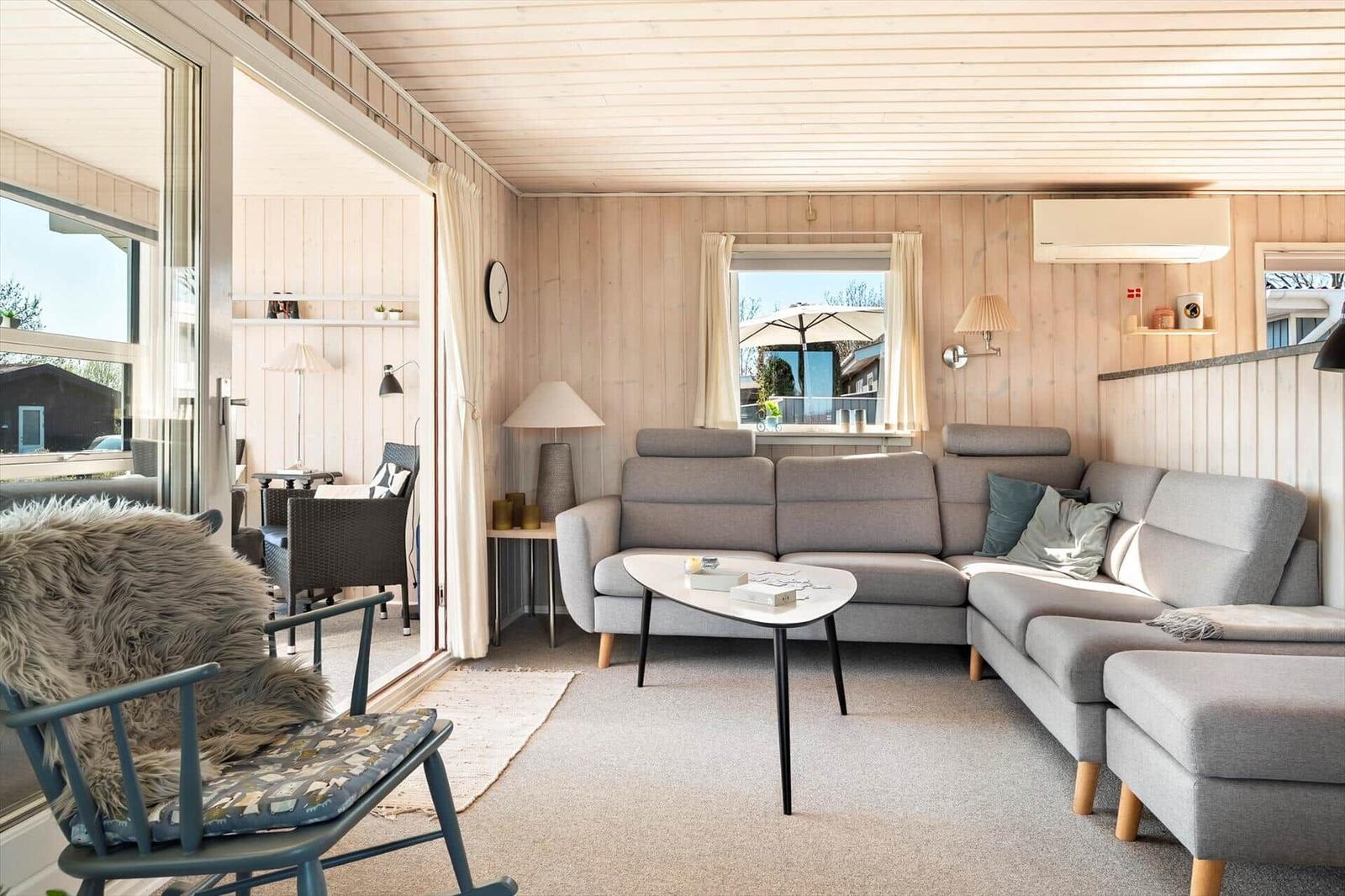 Wohnzimmer mit grauem Sofa, Tisch und Holzwand. Durchgang zur Terrasse mit Sitzgelegenheit.
