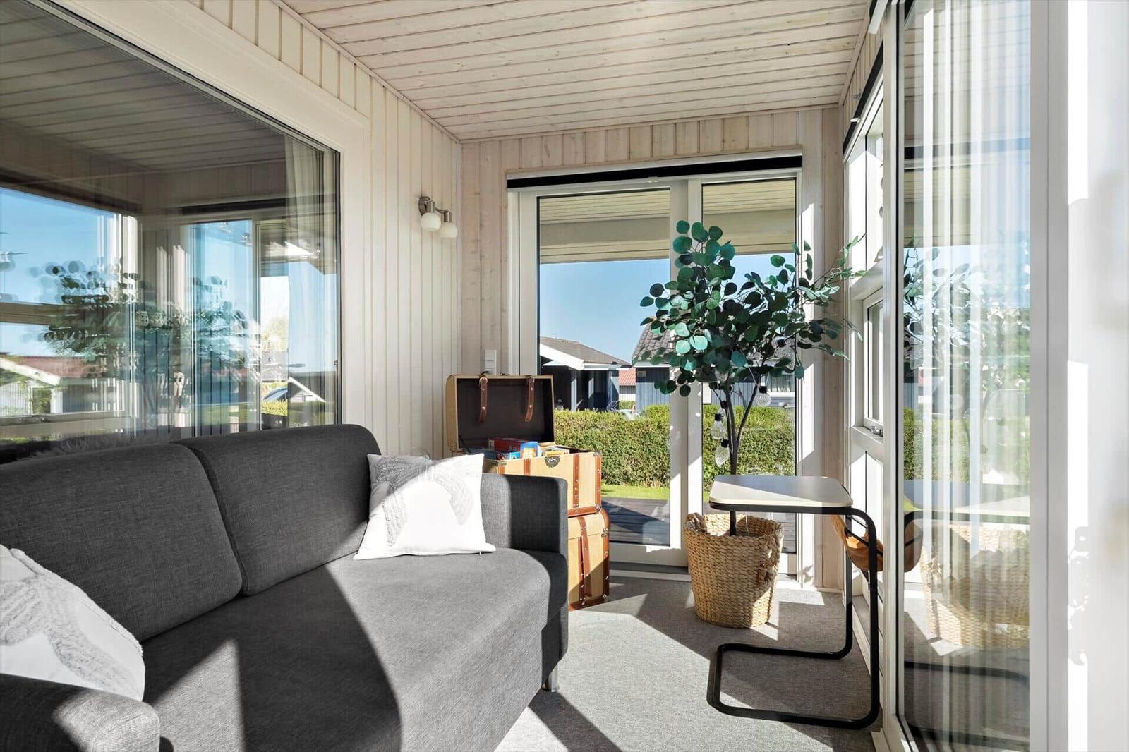 Ein Wohnbereich mit grauem Sofa, Pflanze und Holzwand. Durchgehendes Glas bietet Blick nach draußen.
