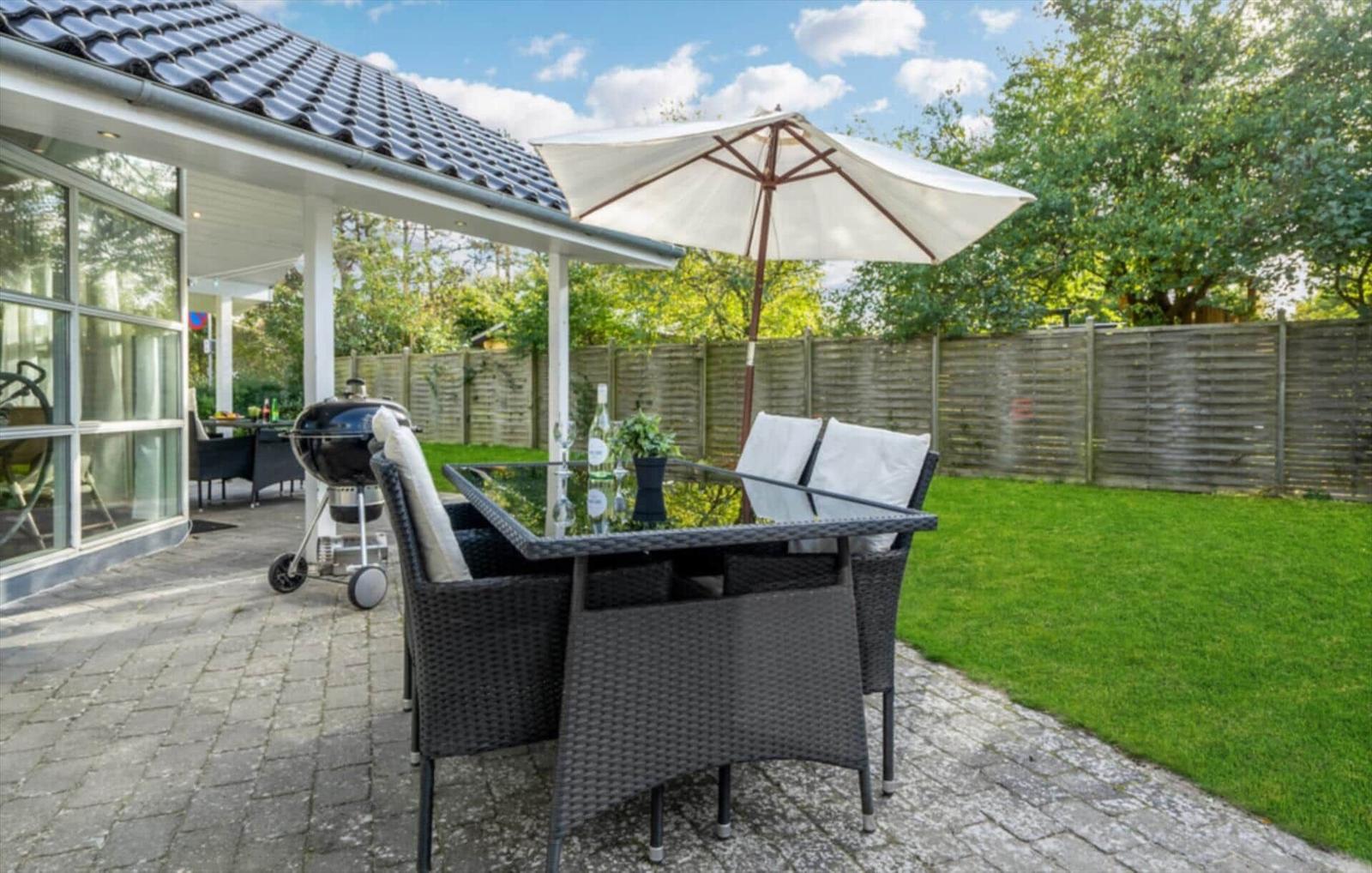 Terrace with table, chairs, umbrella, and grill. Lawn and wooden fence in background.