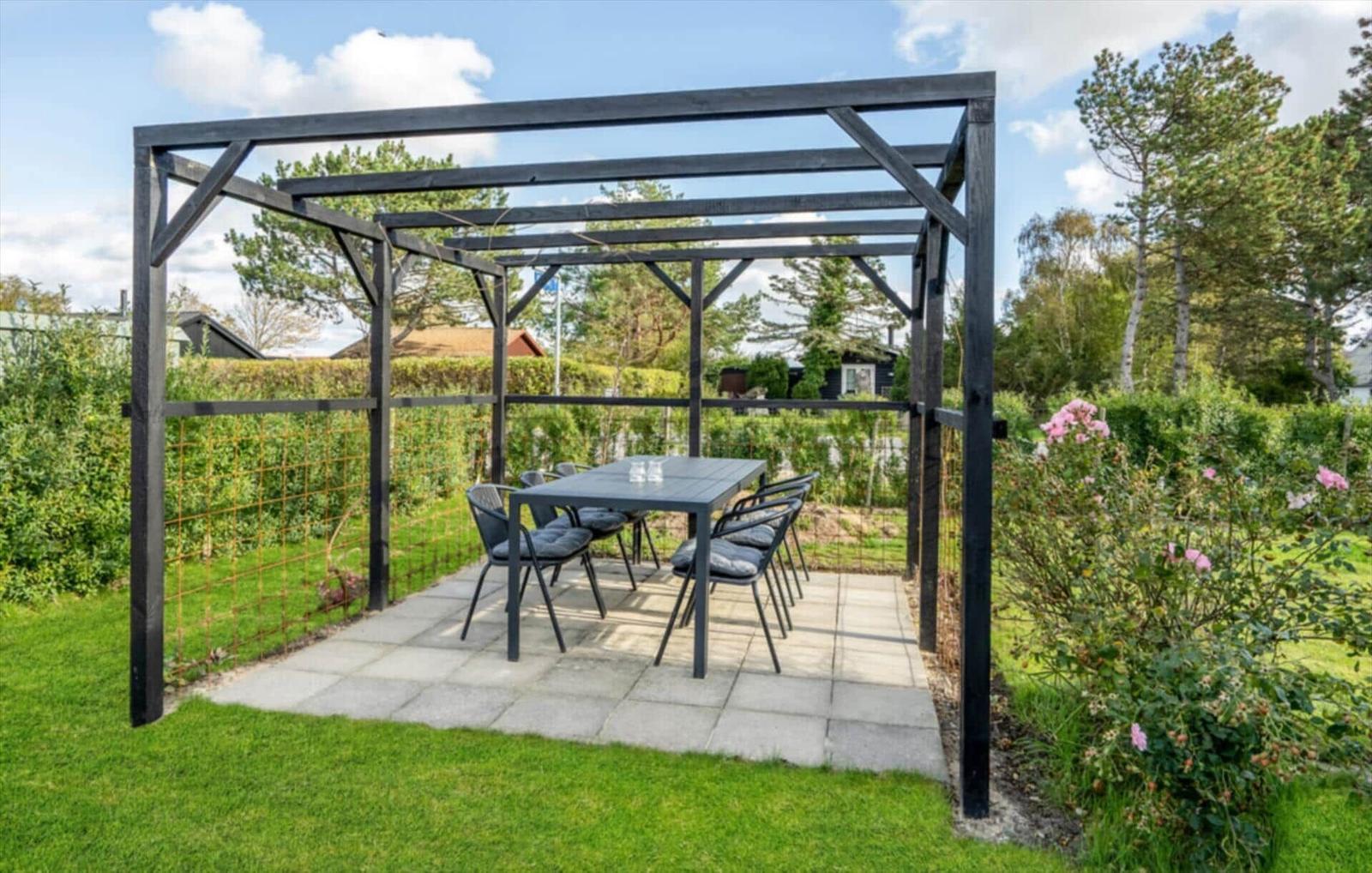Black pergola with table and chairs on stone patio in garden with grass and shrubs.