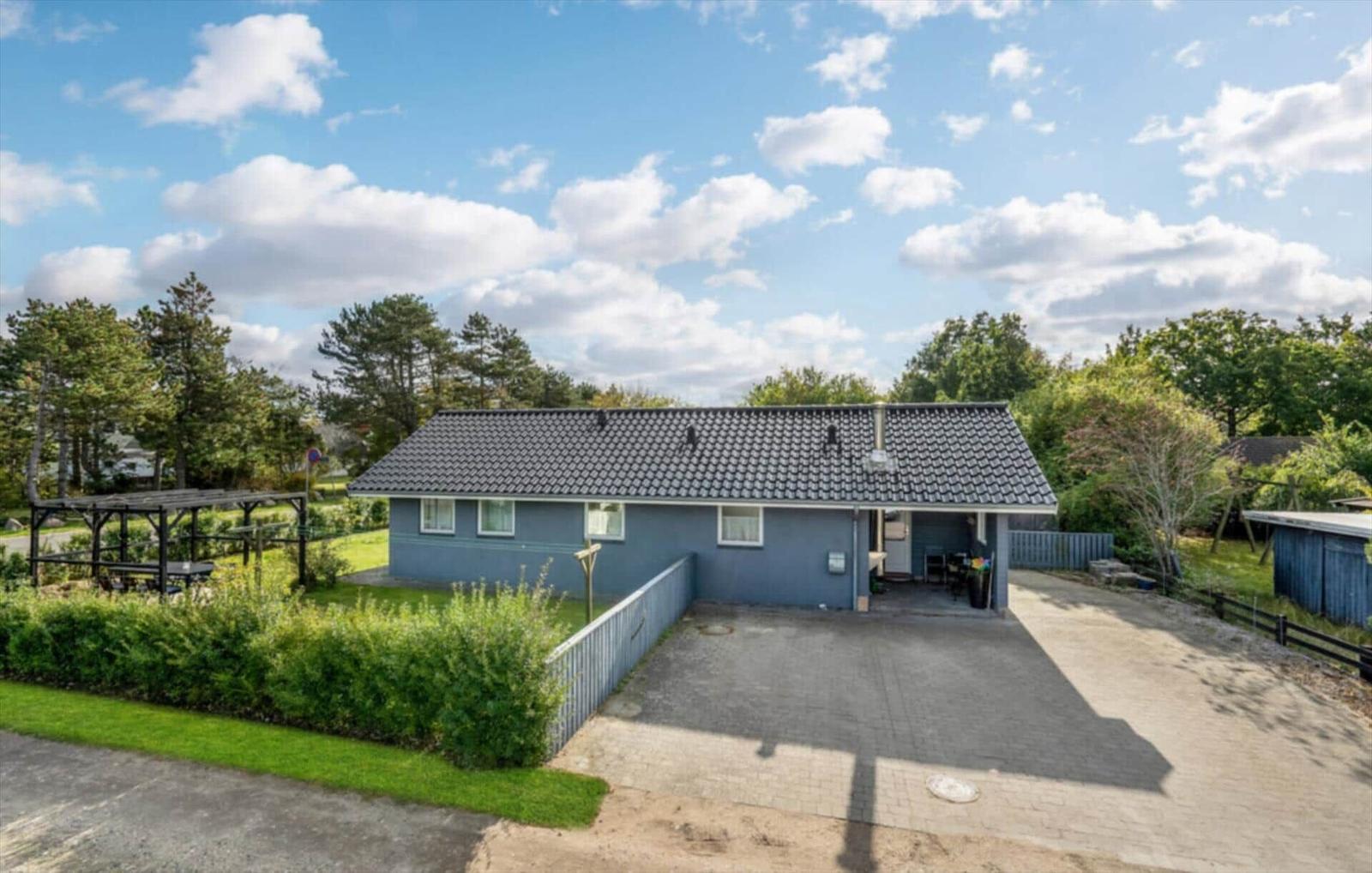 A blue house with a roof, surrounded by greenery and trees under a blue sky with white clouds.