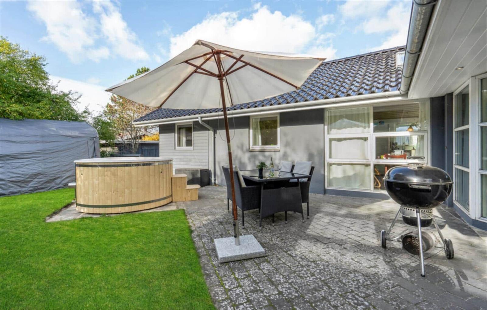 Backyard with hot tub, seating under umbrella, and grill. Lawn and paved patio area.