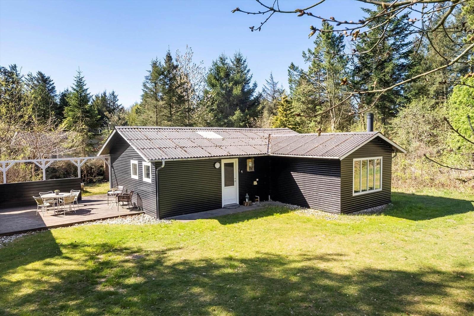 A dark gray wooden house with a terrace and garden. Surrounded by trees and grass.