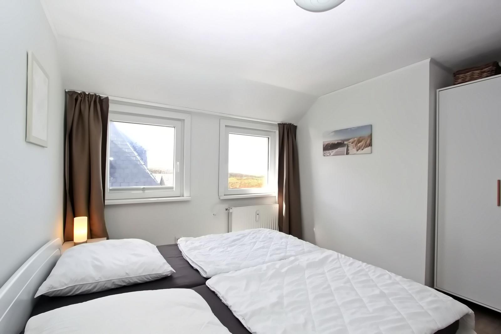 Schlafzimmer mit Doppelbett, zwei Fenstern und Kleiderschrank.