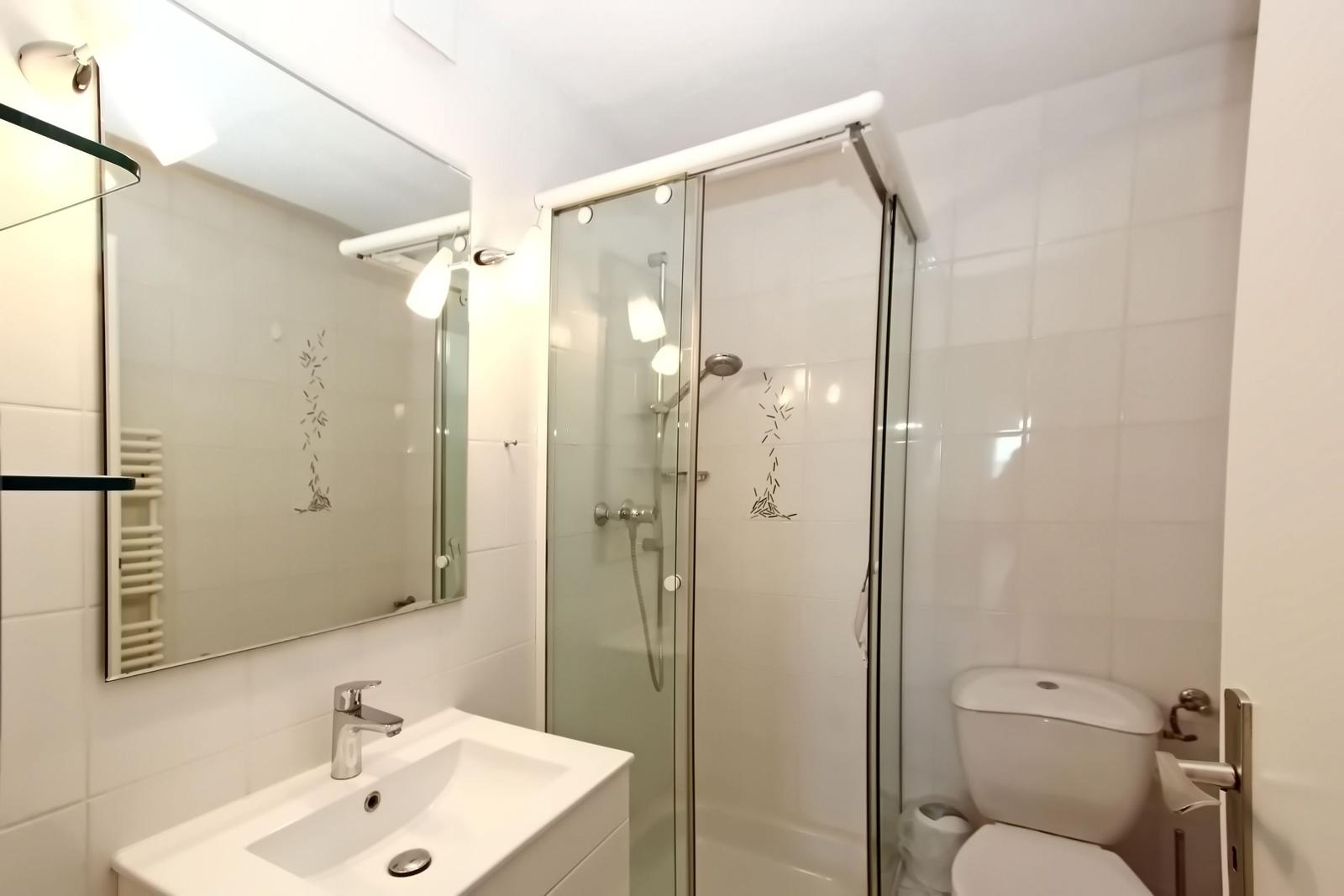 Badezimmer mit Dusche, Waschbecken und Toilette. Weiße Fliesen und Spiegel.