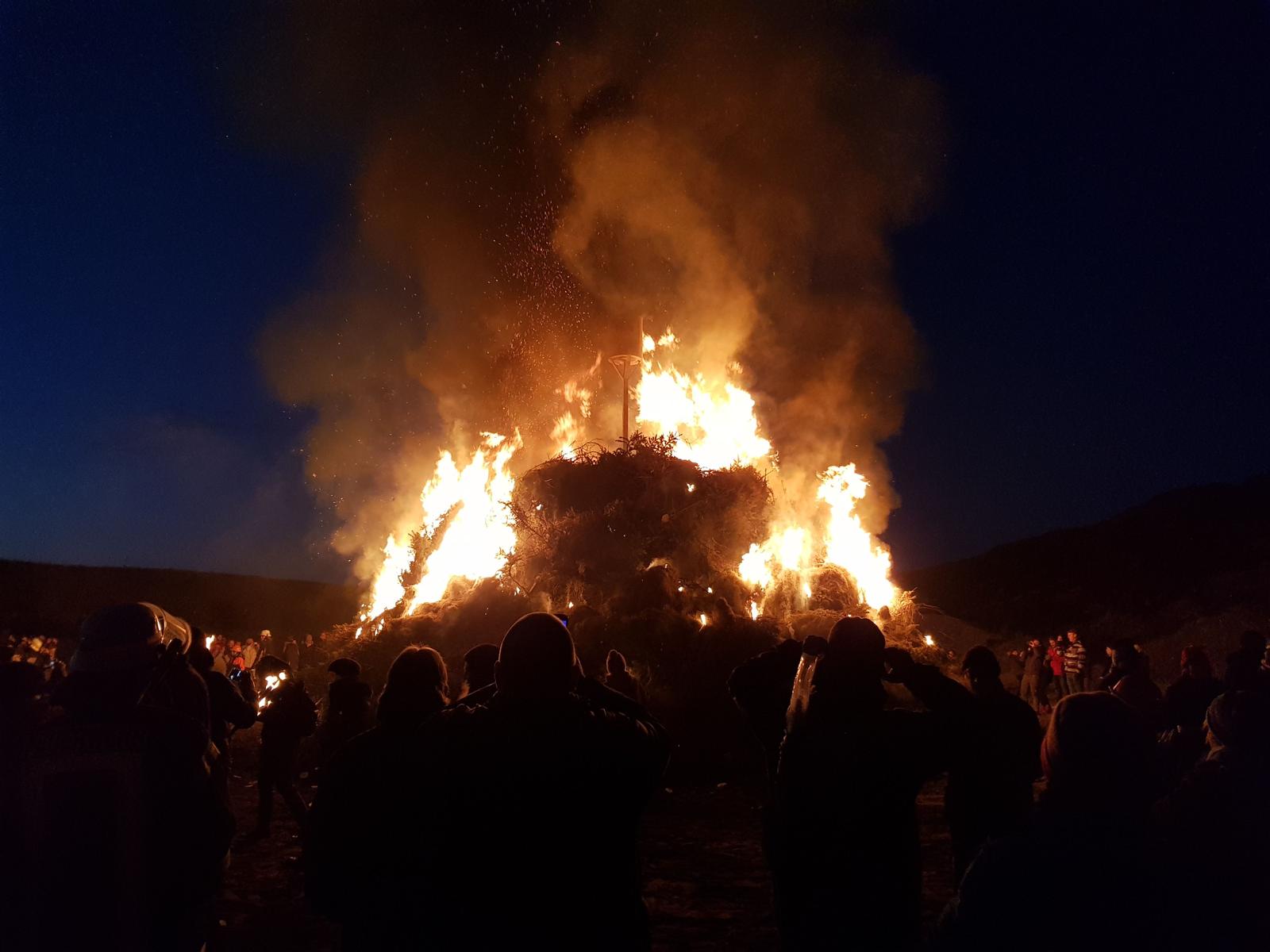Großer Scheiterhaufen brennt bei Nacht. Menschen beobachten das Feuer.