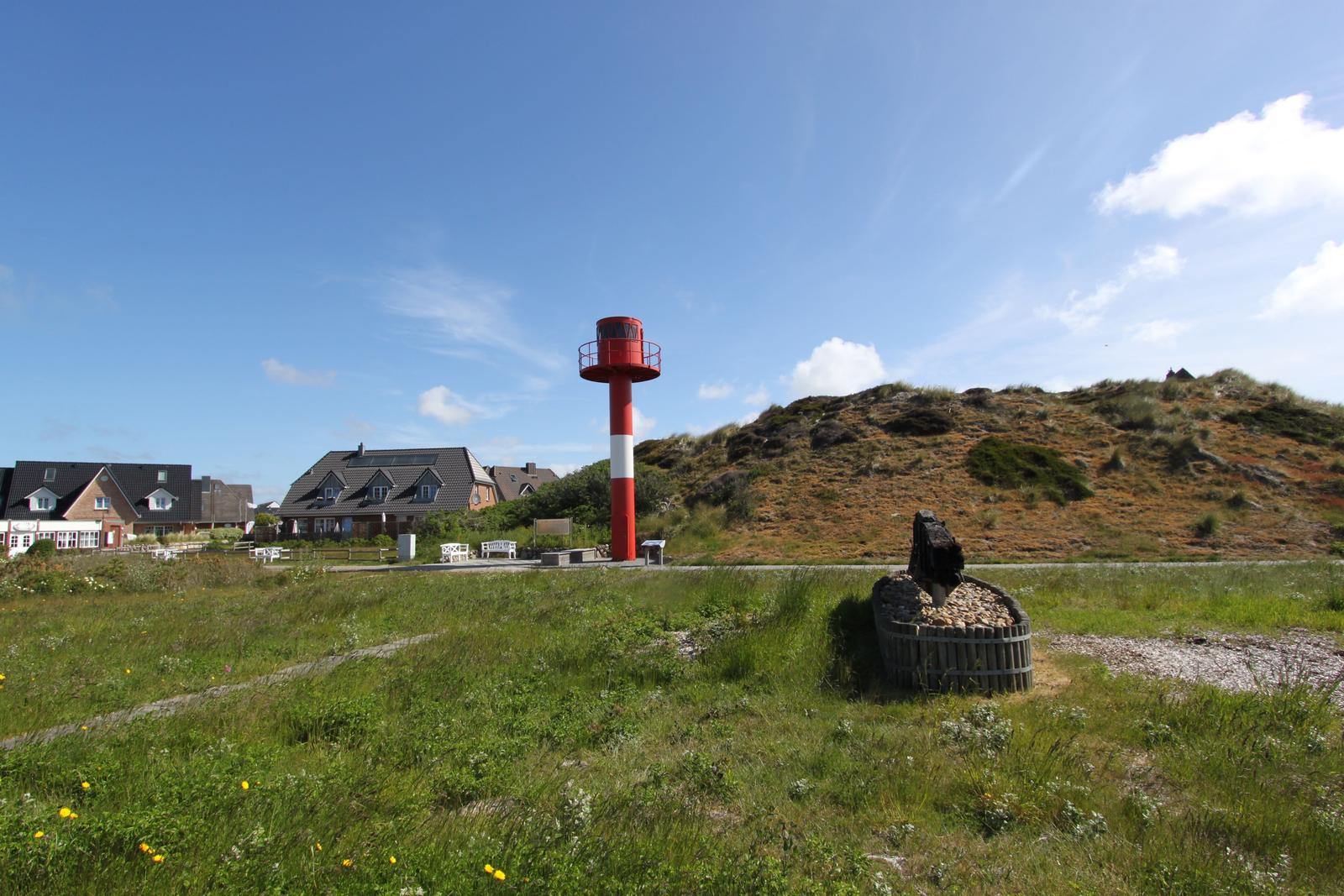 Rotes Leuchtturm mit weißer Markierung, Grasland und Häuser im Hintergrund.