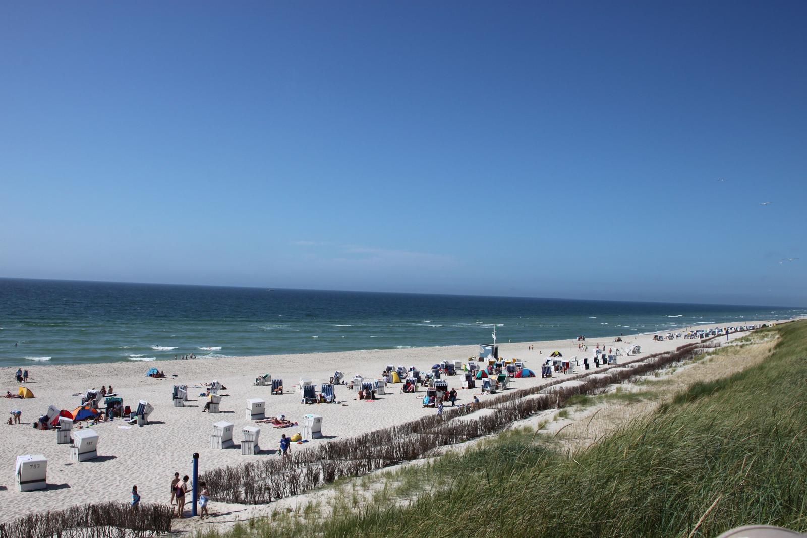 Strand mit weißen Sand, bunten Strandkörben und Menschen unter blauem Himmel.