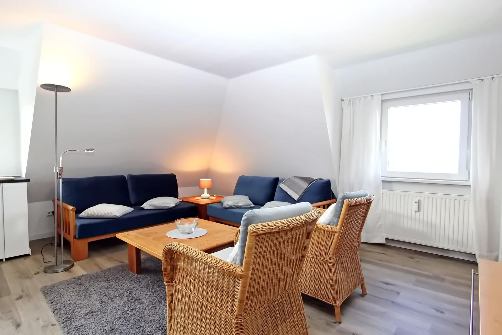 Wohnzimmer mit blauem Sofa, Rattanstühlen und Holztisch. Fenster mit weißen Vorhängen.