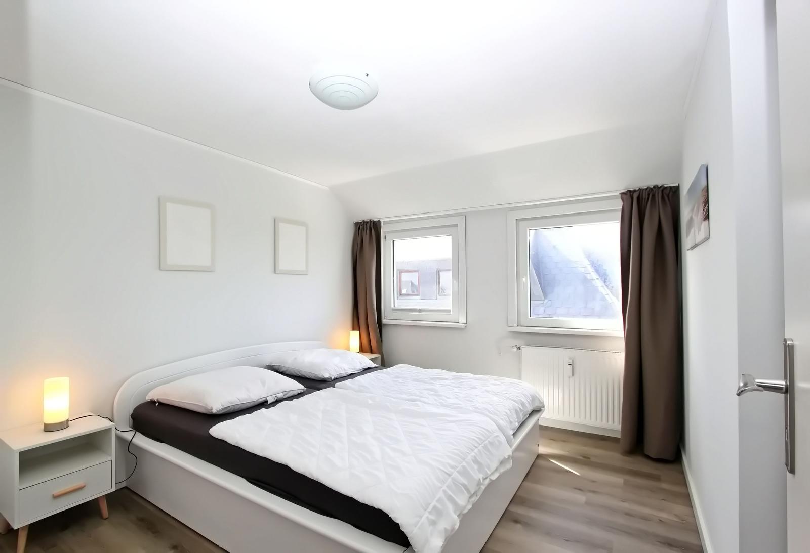 Schlafzimmer mit Doppelbett, zwei Fenstern und Nachttisch mit Lampe.