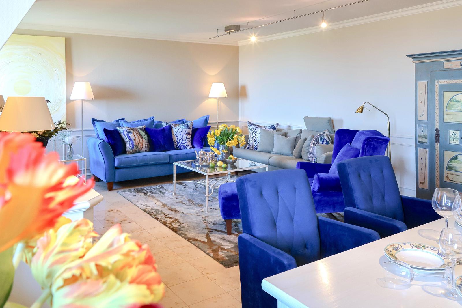 Wohnzimmer mit blauen Sesseln, Sofa und Glas-Tisch. Dekoration mit Blumen und Lampen.
