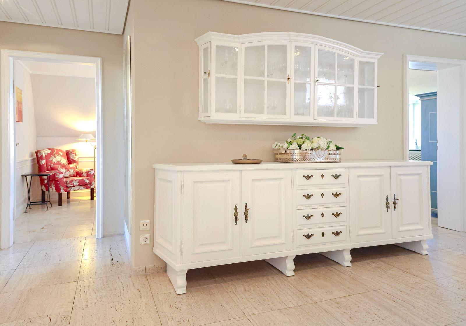 Weißer Sideboard mit Schubladen und darüber hängender Schrank. Im Hintergrund ist ein roter Sessel sichtbar.