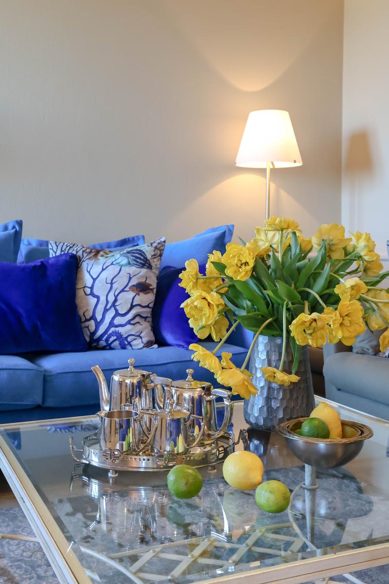 Blauer Sofa mit Kissen, Glas-Tisch mit Teeservice und gelben Tulpen.