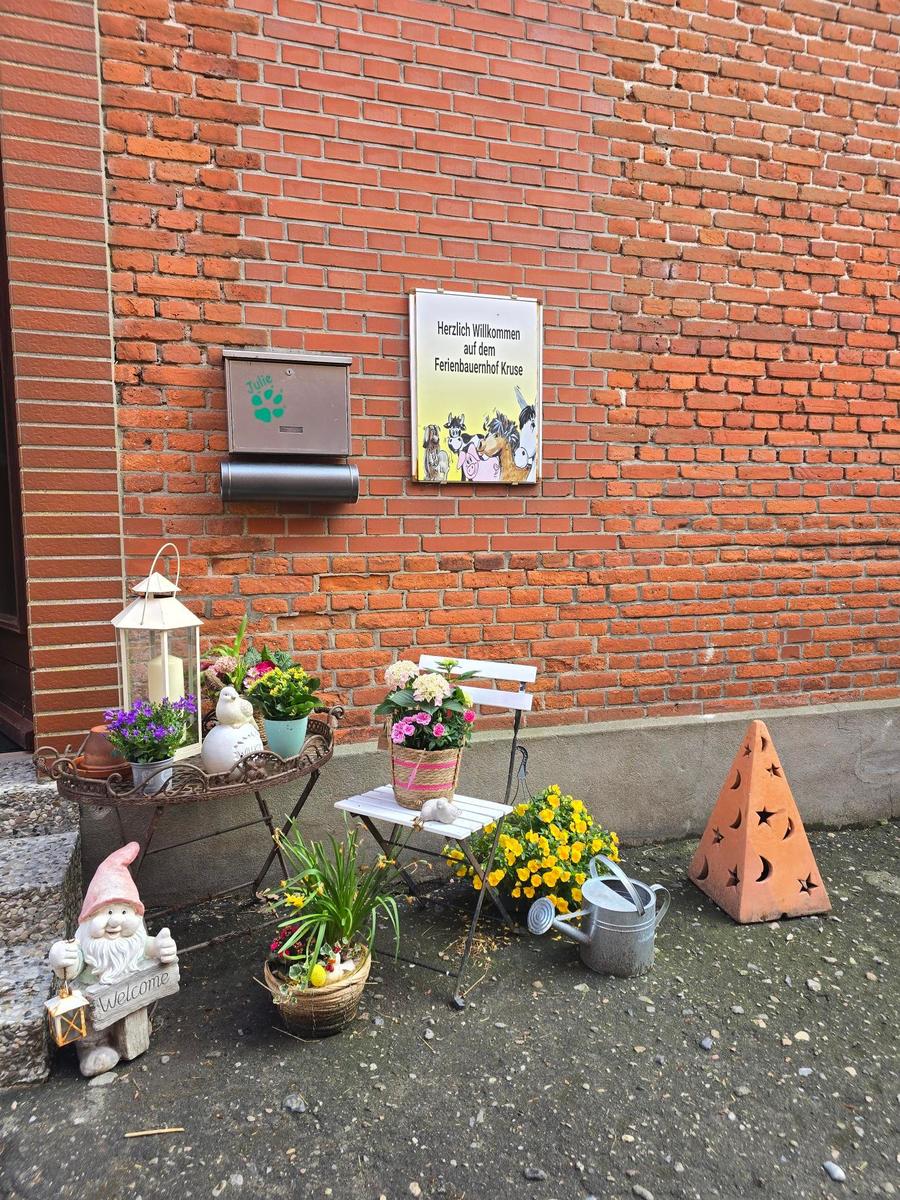 Willkommensschild an Backsteinwand mit Blumen, Stuhl und Gartendekoration.