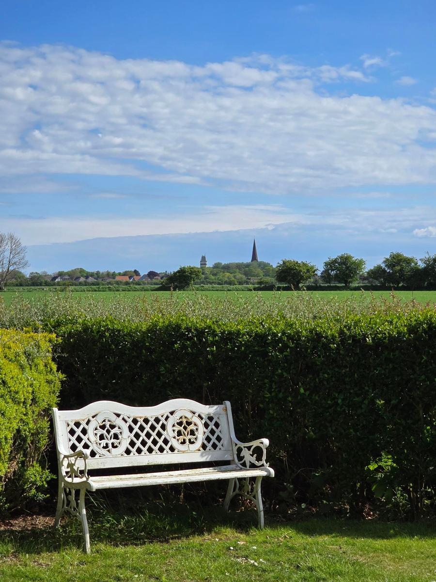 Eine weiße Bank steht im Garten vor einem Heckenzaun. Im Hintergrund sind Felder und ein Dorf mit Kirchturm zu sehen.