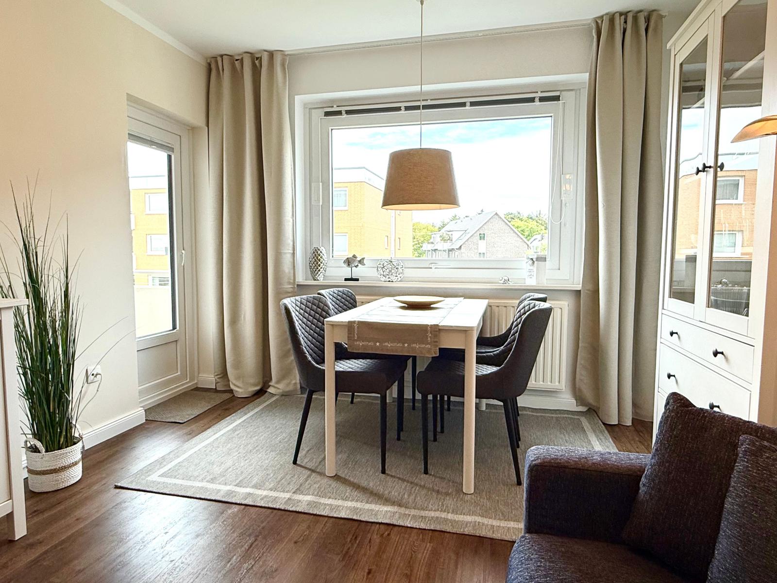 Esszimmer mit Tisch, Stühlen und Fenster. Vorhang, Pflanze und Schrank sichtbar.