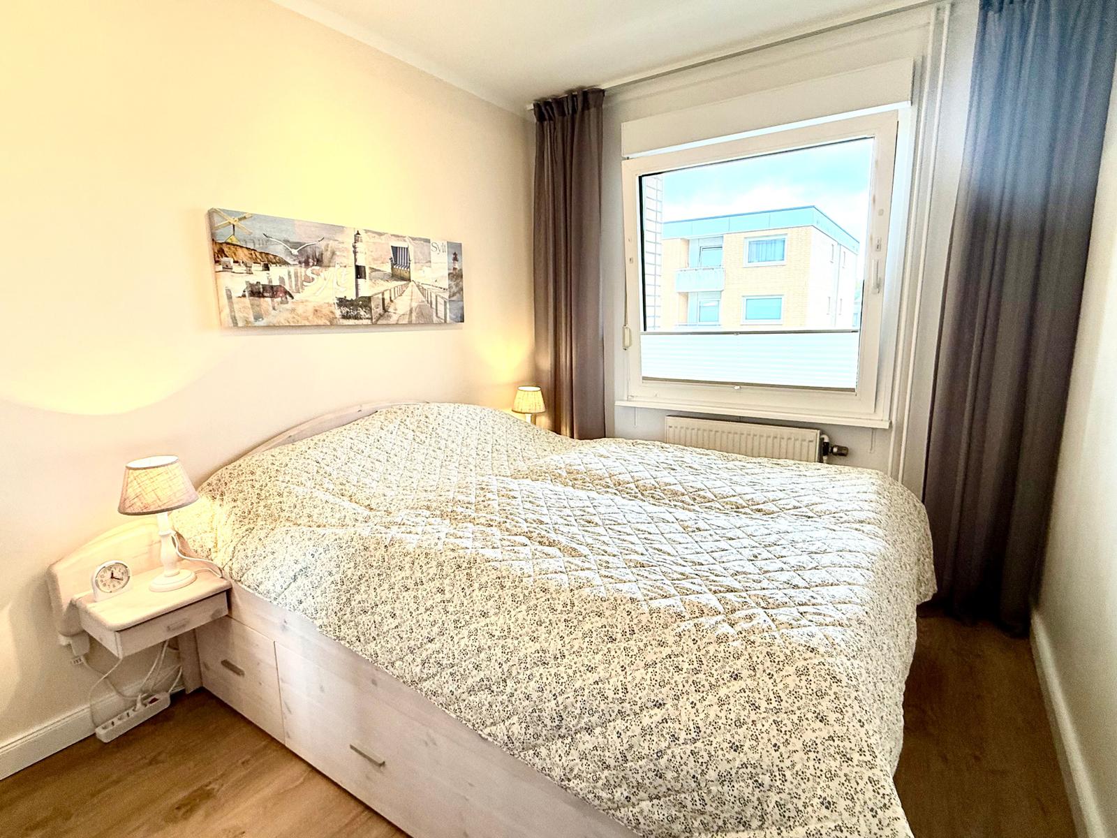 Schlafzimmer mit Doppelbett, Nachttisch und Fensterblick auf Gebäude.