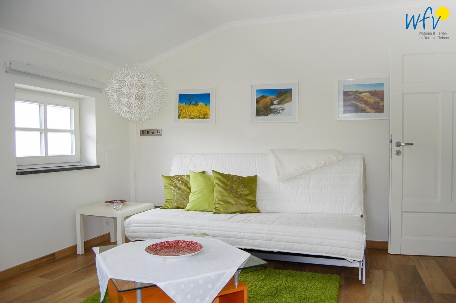 Ein Wohnzimmer mit weißem Sofa, grünen Kissen und Holzboden. An der Wand hängen drei Bilder.