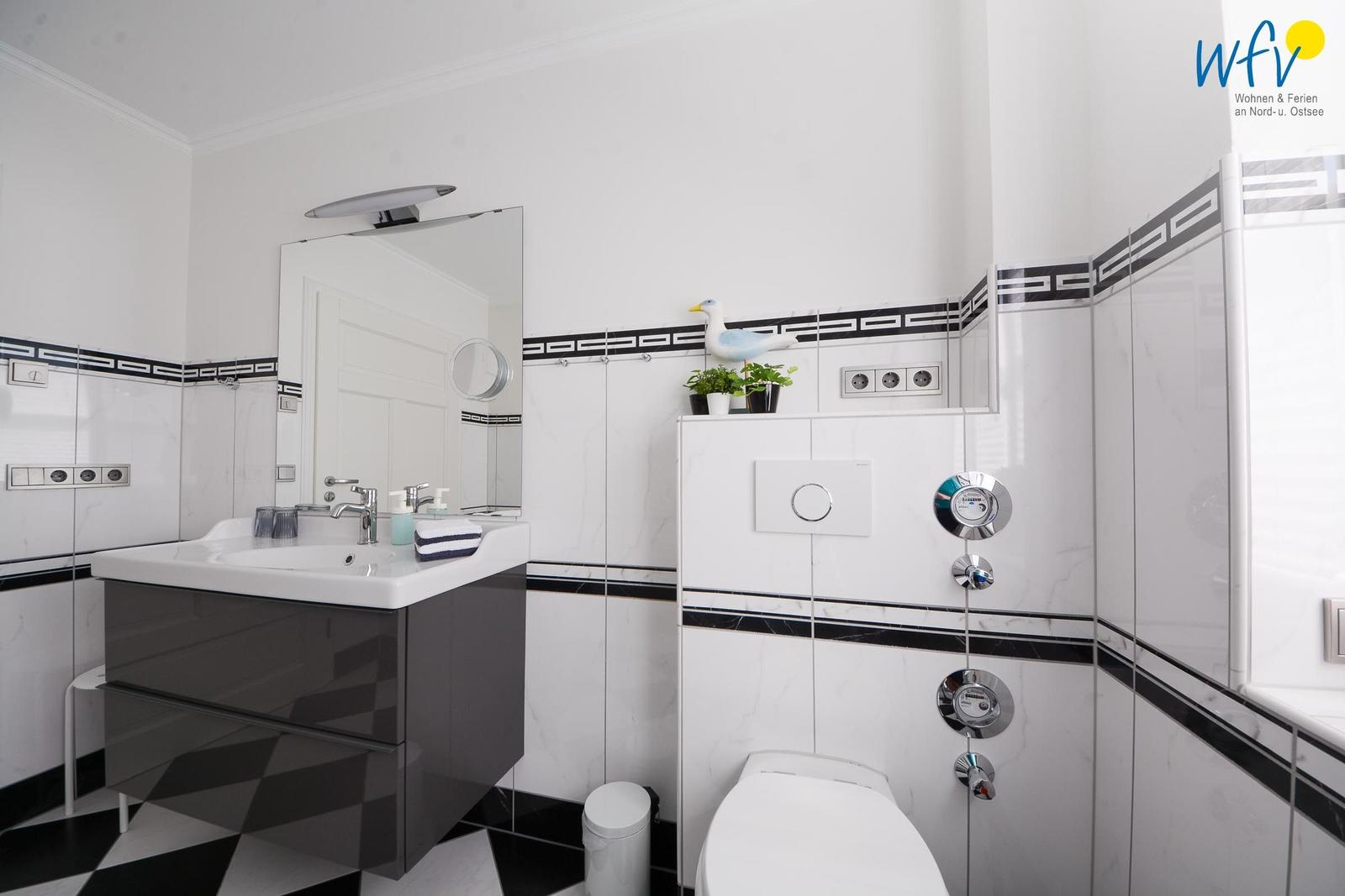 Badezimmer mit Waschbecken, Spiegel, Toilette und Dusche. Weiße Fliesen mit schwarzen Akzenten.
