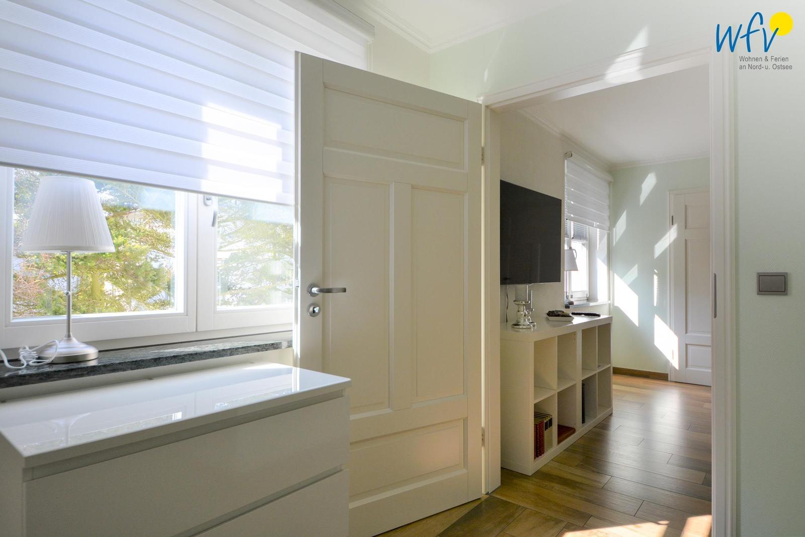 Schlafzimmer mit Fenster, Schrank und Tür zum Flur. Fernseher auf Holzregal.
