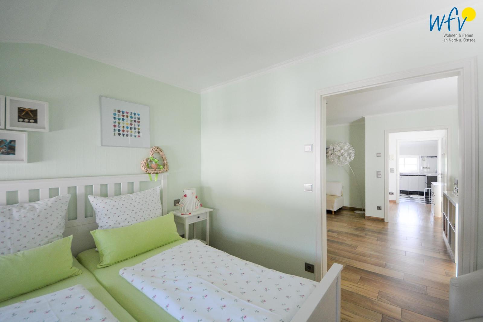 Schlafzimmer mit Doppelbett, grünen Kissen und Bildern an der Wand. Durchgang zu Wohn- und Küchenbereich.