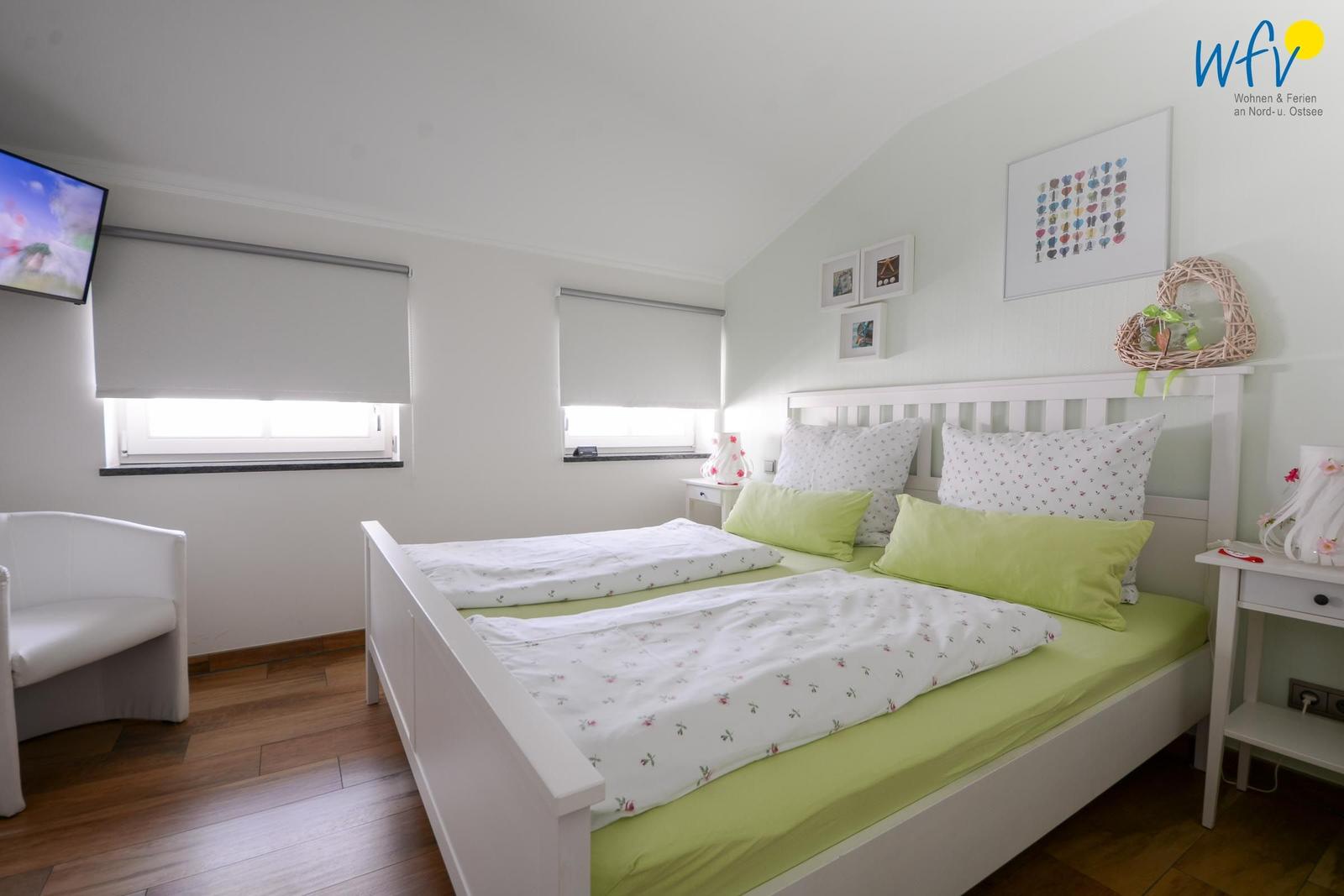 Ein Schlafzimmer mit Doppelbett, zwei Fenstern und einer TV-Wand