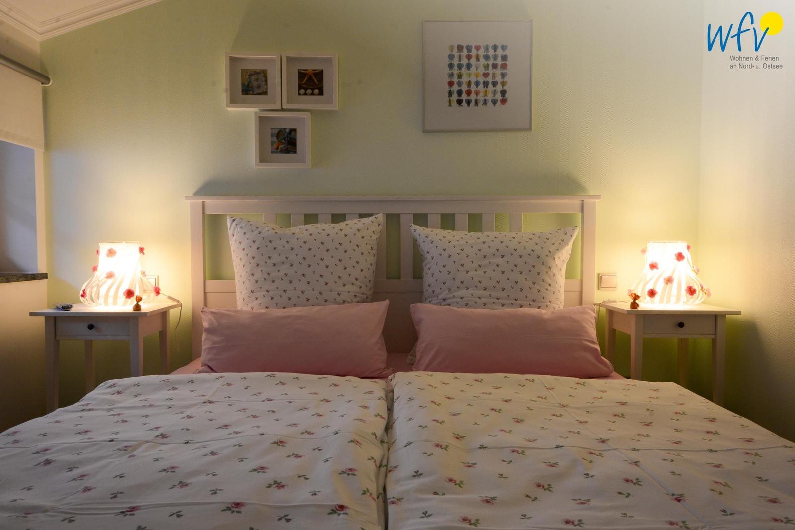 Schlafzimmer mit Doppelbett, zwei Nachttischen und Lampen. Wandbilder über dem Bett.