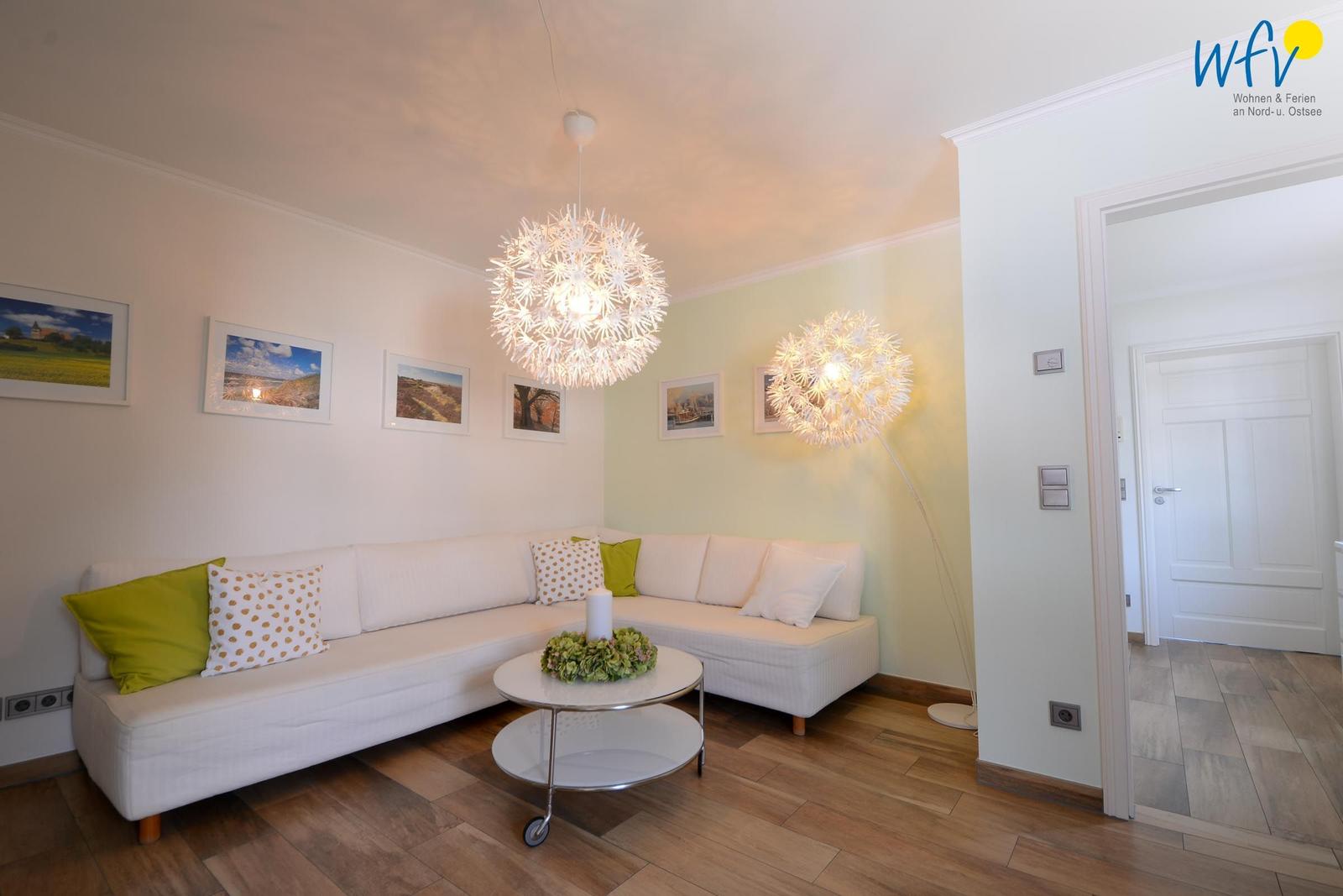 Wohnzimmer mit weißem Sofa, grünen Kissen und runden Tisch. Zwei Doldenlampen hängen von der Decke.