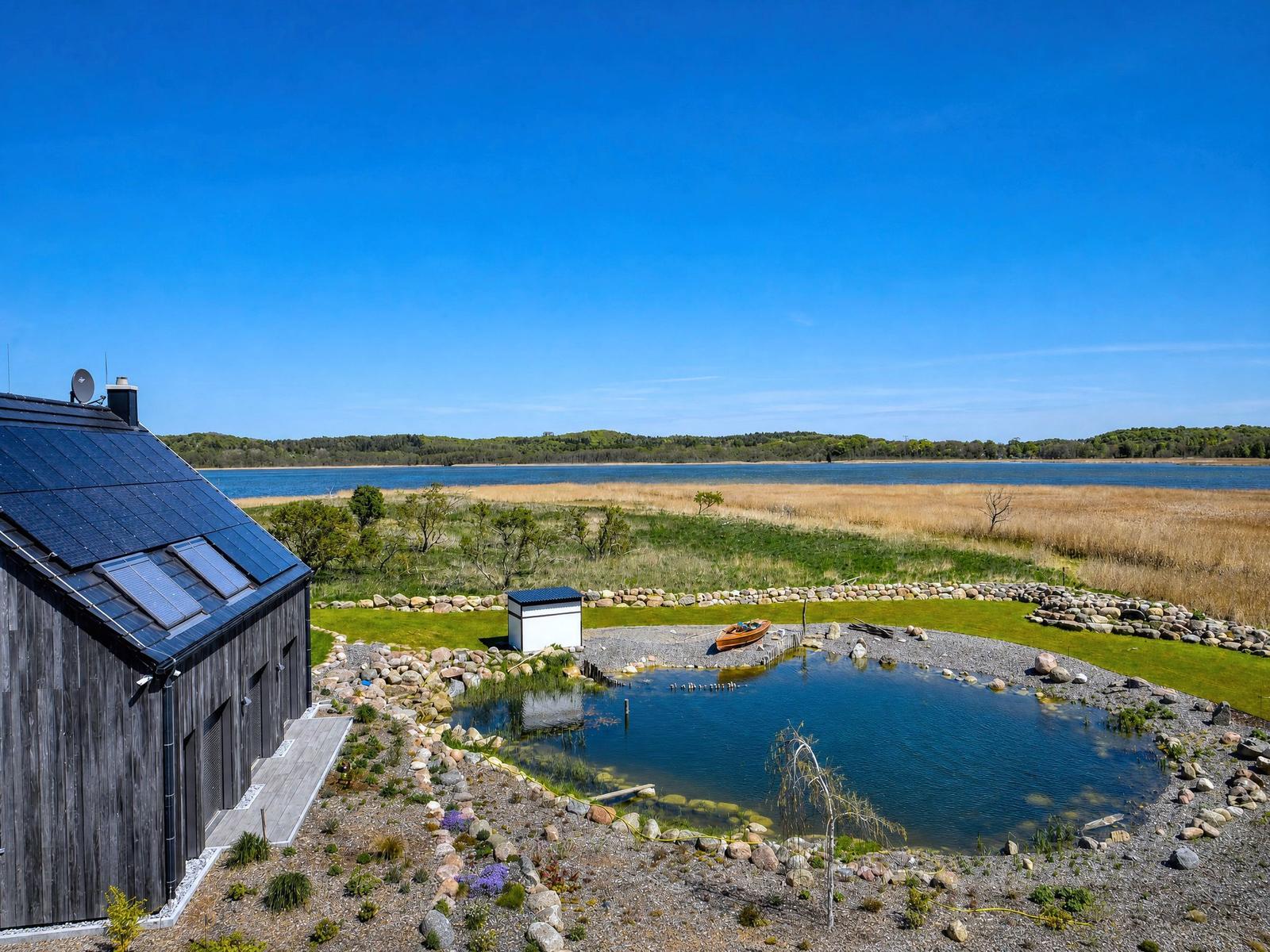 House with solar panels, small pond, and boat. View of calm water and green land.