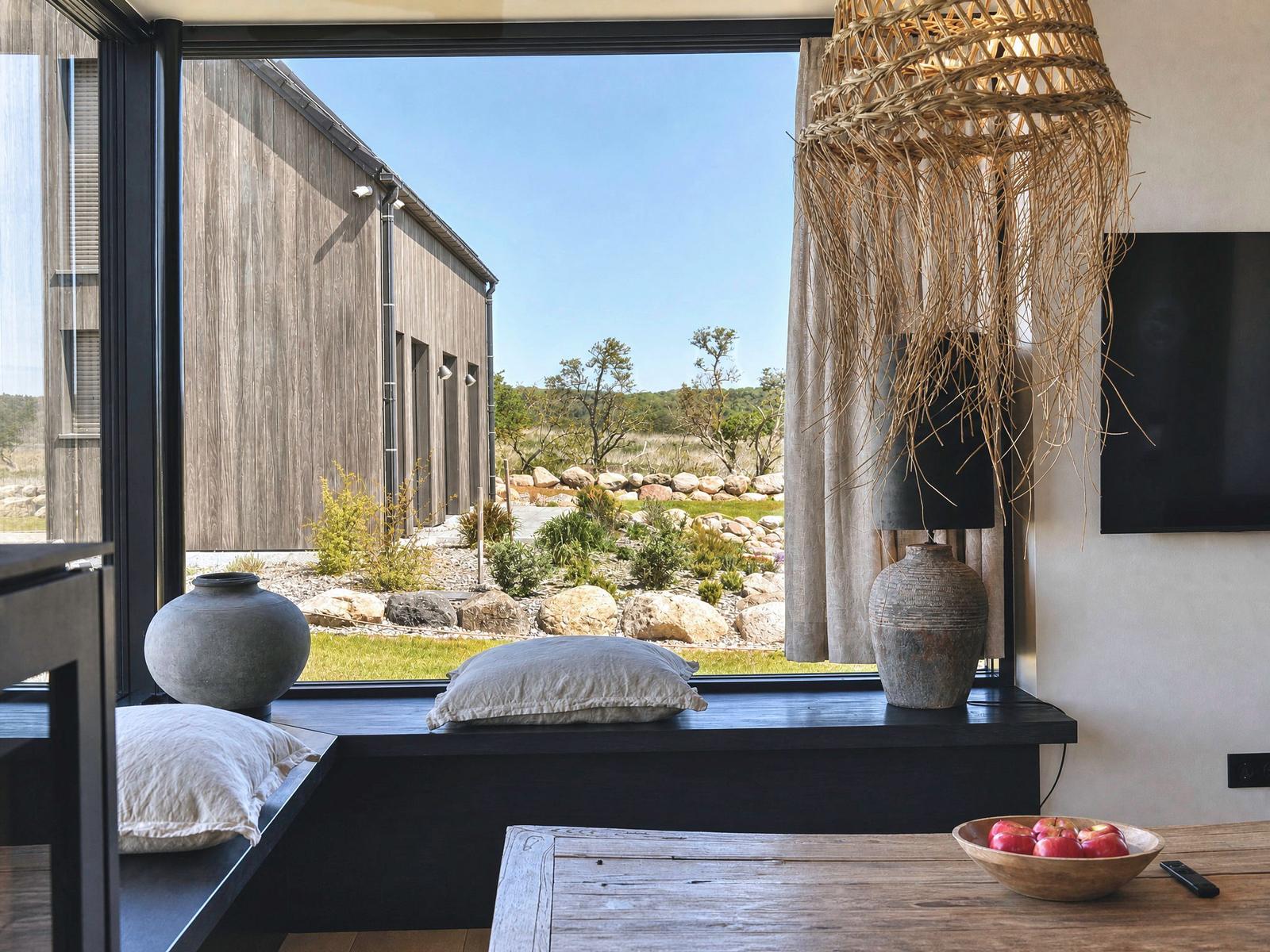 Large window overlooking garden and wooden house. Seat with cushions, table with apples.