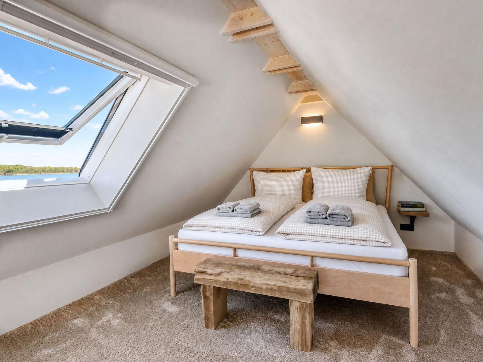 Attic room with bed, wooden bench, and skylight overlooking water.
